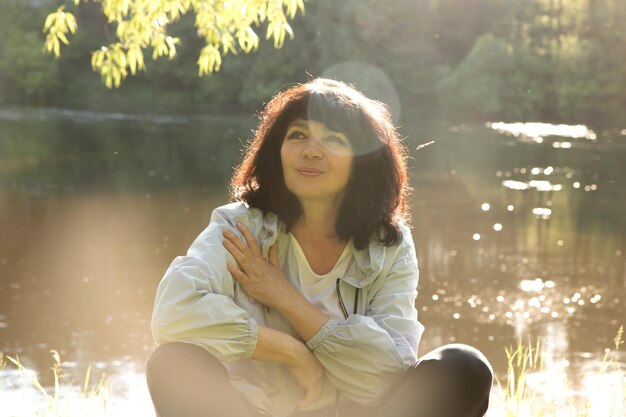 Sommerporträt Eine Frau mit Sonnenblendung im Gesicht in der untergehenden Sonne sitzt am Flussufer