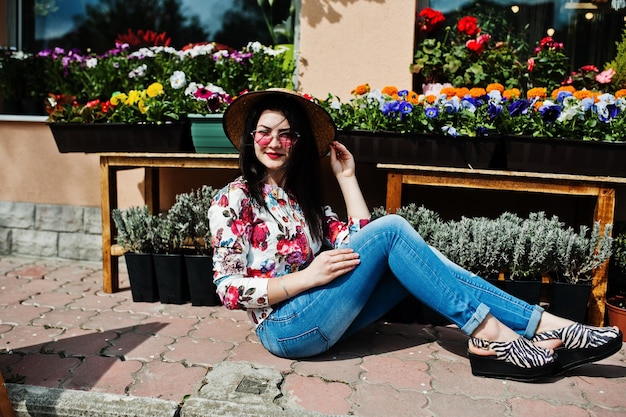 Sommerporträt eines brünetten Mädchens mit rosa Brille und Hut gegen Blumenladen