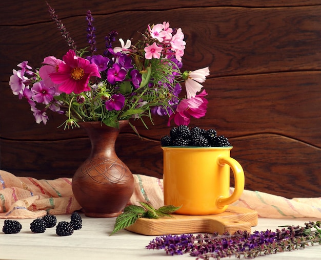 Sommerstillleben Ein Strauß Sommerblumen in einer Tonvase Ein gelber Becher Brombeeren blüht in der Nähe von Holzhintergrund Platz für Text