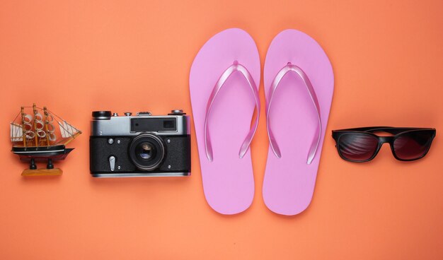 Sommerstillleben. Strandzubehör. Modische rosa Flipflops, Tasche, Retro-Kamera, Sonnenbrille, Schiff auf Korallenpapierhintergrund.