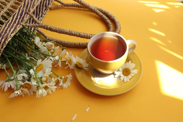 Sommerstimmung eine gelbe Tasse Tee in der Nähe eines Korbes mit einem Strauß Gänseblümchen in der Sonne auf gelbem Hintergrund Platz für Text