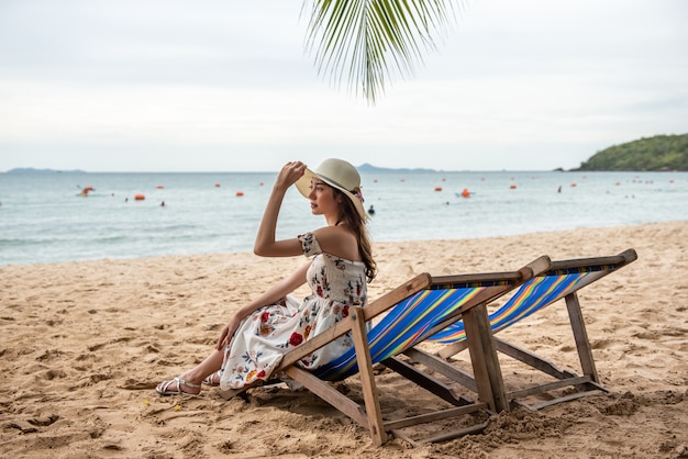 Sommerstrandferien-Feiertagsreisekonzept, glückliche junge Asiatin mit dem Hut, der sich oben auf Strandstuhl und angehobenen Händen entspannt.