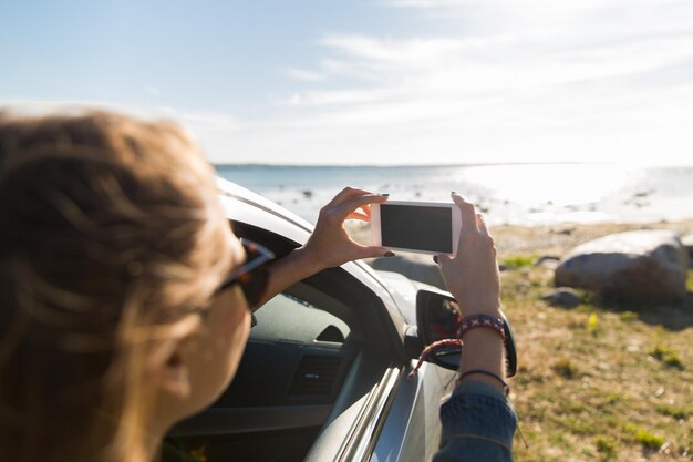 Sommerurlaub, Feiertage, Reisen, Roadtrip und Menschenkonzept – glückliches Teenager-Mädchen oder junge Frau im Auto, die mit dem Smartphone das Meer fotografieren