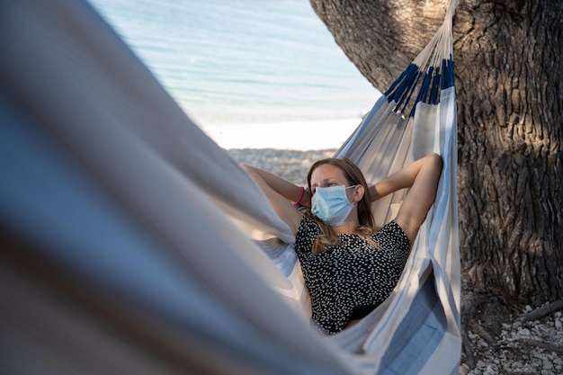 Sommerurlaub während der Coronavirus-Pandemie junge Frau, die in einer Hängematte am Strand liegt und eine schützende medizinische Gesichtsmaske trägt