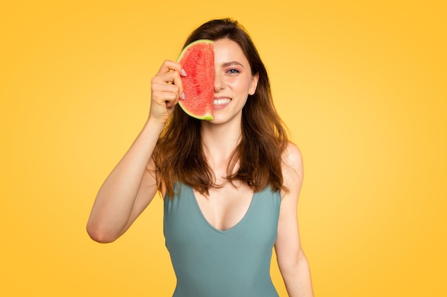 Sommerzeitkonzept hübsche Frau, die die Hälfte der roten Wassermelone in der Nähe des Gesichts hält und über Gelb posiert
