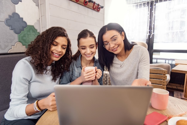 Somos los mejores. Agradables tres estudiantes joviales sentados a la mesa y mirando la pantalla mientras beben té y usan teléfonos inteligentes