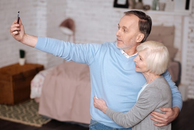 Somos modernos. Retrato de homem idoso bonito abraçando sua esposa e tomando selfie em casa.