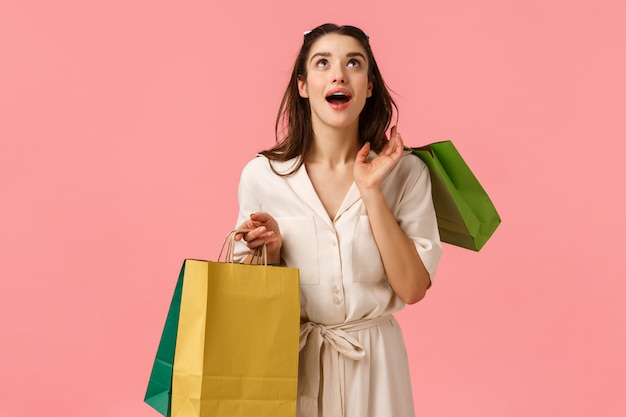 Soñadora, alegre, divertida y alegre chica morena vestida, sosteniendo bolsas de compras, mirando sorprendida y asombrada, sonriendo maravillada, sintiéndose feliz paseando por los centros comerciales de la ciudad