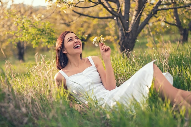 Soñadora alegre en vestido blanco sosteniendo flores y riendo mientras está acostado sobre la hierba verde en el jardín de primavera en un día soleado