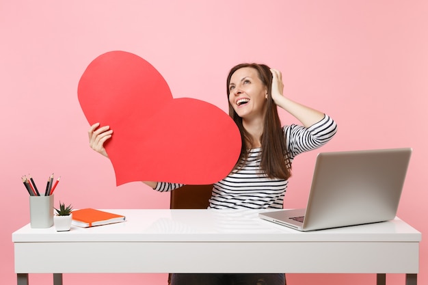 Soñadora feliz niña mirando hacia arriba sosteniendo rojo corazón en blanco vacío sentarse a trabajar en el escritorio blanco con ordenador portátil aislado sobre fondo rosa pastel. Concepto de carrera empresarial de logro. Copie el espacio para publicidad.