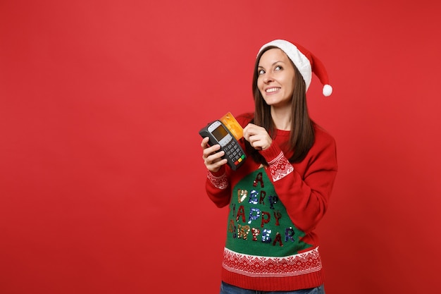 Soñadora joven Santa mantenga terminal inalámbrico de pago bancario moderno para procesar y adquirir pagos con tarjeta de crédito, tarjeta aislada sobre fondo rojo. Feliz año nuevo 2019 celebración concepto de fiesta navideña.