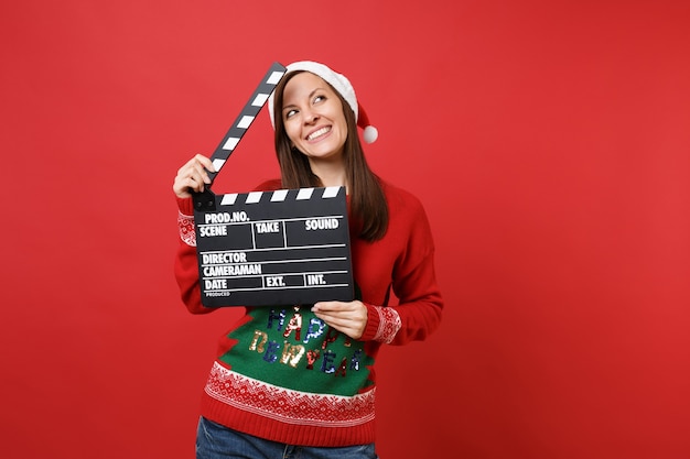 Soñadora joven Santa con sombrero de Navidad mirando hacia arriba, sostenga la clásica claqueta de cine negra aislada sobre fondo rojo. Feliz año nuevo 2019 celebración concepto de fiesta navideña. Simulacros de espacio de copia.