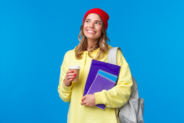 Soñadora linda chica universitaria que contempla el soleado día de primavera en su camino a las clases, sosteniendo café para llevar y cuadernos, sonriendo mientras mira hacia arriba, llevar mochila, de pie