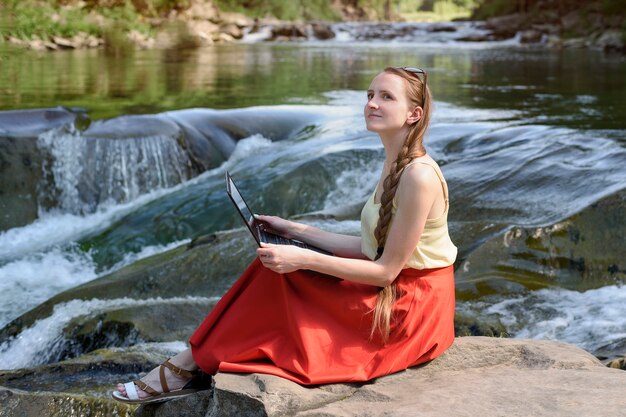 Soñando con una hermosa chica de pelo largo con una falda roja sentada con una computadora portátil en una piedra con el telón de fondo de una cascada de un río de montaña. Concepto de autónomo. Trabaja en la naturaleza.