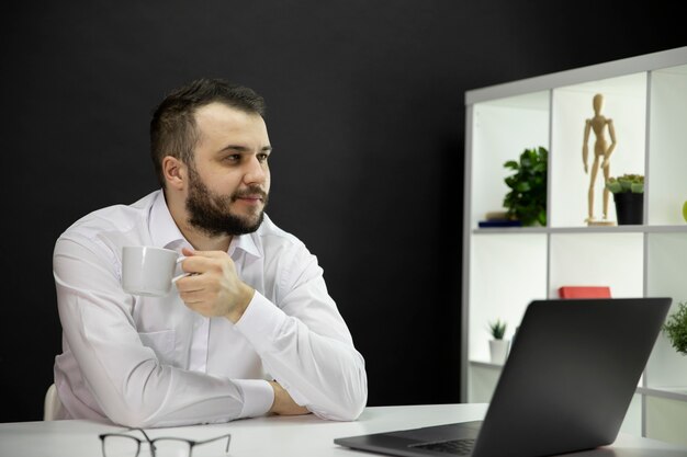 Soñando hombre guapo con una taza de café sentado a la mesa frente a la computadora portátil