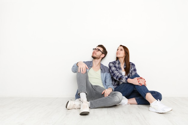 Foto soñando juntos, la pareja mira hacia arriba, copie el espacio. hombre joven y mujer sentada en el suelo en casa junto a la pared blanca en blanco