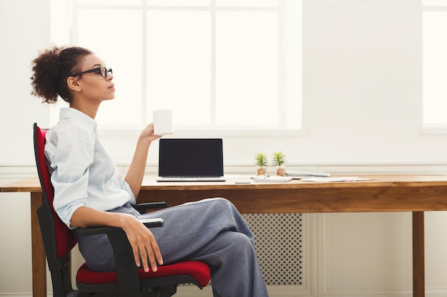 Soñando con vacaciones. Mujer de negocios relajada sentada en el lugar de trabajo en la oficina moderna con una taza de café, copie el espacio, vista lateral
