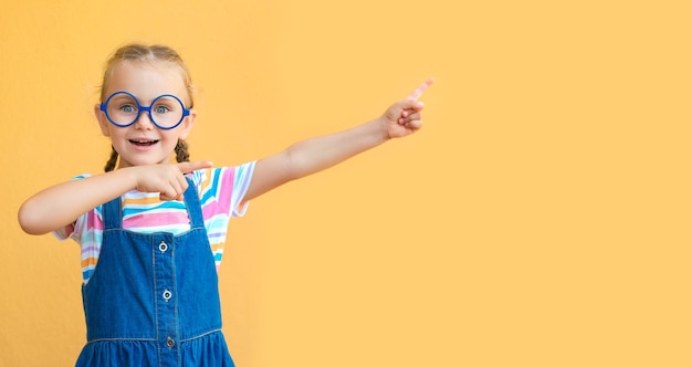 Sonderangebot Lächelndes, entzückendes Mädchen mit Brille, das auf den Kopierbereich über gelbem Hintergrund zeigt Farbige gestreifte Kleidung