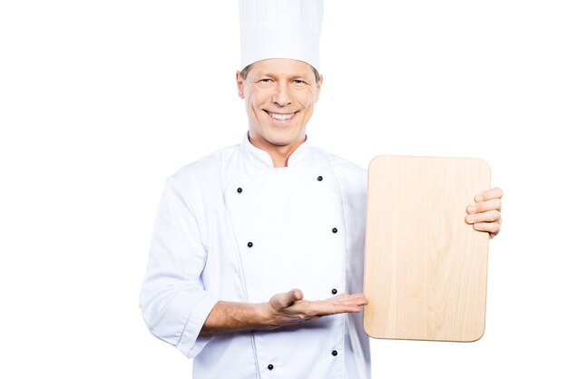 Sonderangebot vom Küchenchef. Selbstbewusster reifer Koch in weißer Uniform, der ein Holzschneidebrett hält und mit einem Lächeln darauf zeigt, während er vor weißem Hintergrund steht