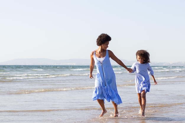 Sonhadora mãe e filha de férias. Família afro-americana em lindos vestidos andando na praia, se divertindo, correndo. Família, viagem, conceito de paternidade