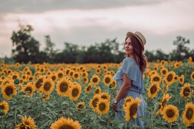 Sonhando com uma jovem mulher de vestido azul e chapéu em um campo de girassóis no verão, vista do lado dela. Olhando para o lado. copie o espaço