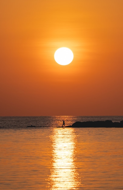 Sonne über dem Meer am orangefarbenen Himmel. Fischer mit Angelrute auf dem Felsen.