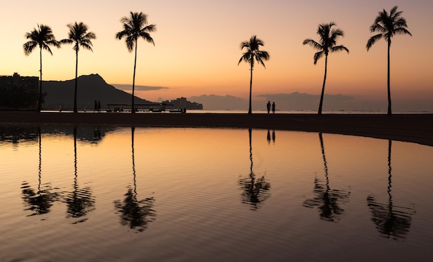Sonnenaufgang in Waikiki mit Diamond Head, der sich im ruhigen Wasser des Teichs widerspiegelt, während Wanderer am Strand entlang gehen