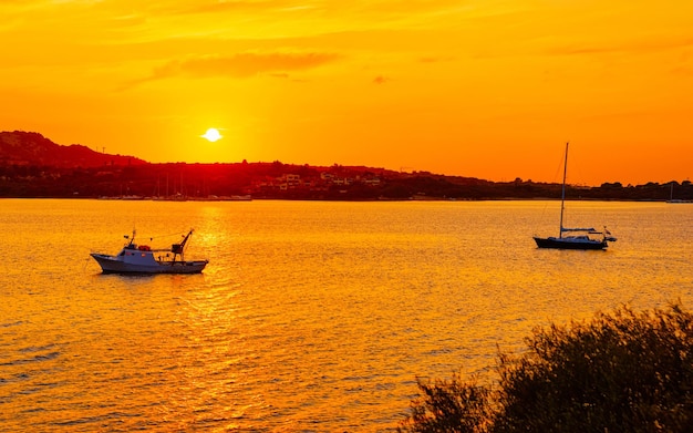 Sonnenaufgang oder Sonnenuntergang mit Yachten in Porto Rotondo an der Costa Smeralda am Mittelmeer auf der italienischen Insel Sardinien. Boot in Sardinien im Sommer. Landschaft der Provinz Olbia. Gemischte Medien.