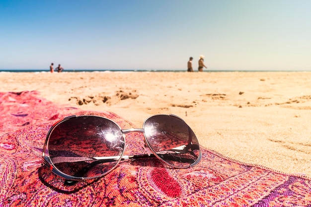 Sonnenbrille auf einem Strandtuch im Hintergrund das Ufer mit Passanten
