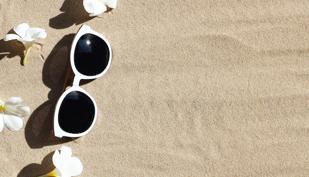 Sonnenbrille mit weißer Plumeriablume auf Sand. Sommer Hintergrundkonzept.