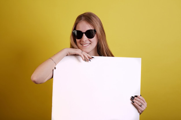 Sonnenbrilleningermädchen versteckt sich für weiße Malerei auf gelbem Hintergrund