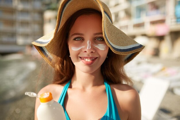 Sonnencreme Frau, die Sonnencreme auf das Gesicht aufträgt Schönes, glückliches, süßes Mädchen legt Sonnencreme aus einer Plastikbehälterflasche auf ihre Nase und Wangen Frau in Strohhut und Bikini am Strand