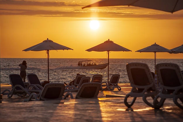 Sonnenschirme und Sonnenliegen schaffen eine Sonnenuntergangssilhouette am Strand von Dominicus in der Dominikanischen Republik mit einem Motorboot im Meer