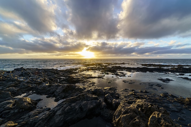 Sonnenuntergang an der Seeküste mit großen Felsen, die von den Wellen gewaschen werden, Himmel mit Wolken in goldenen Tönen und Reflexionen im Wasser zwischen den Felsen. Spanien. Europa.