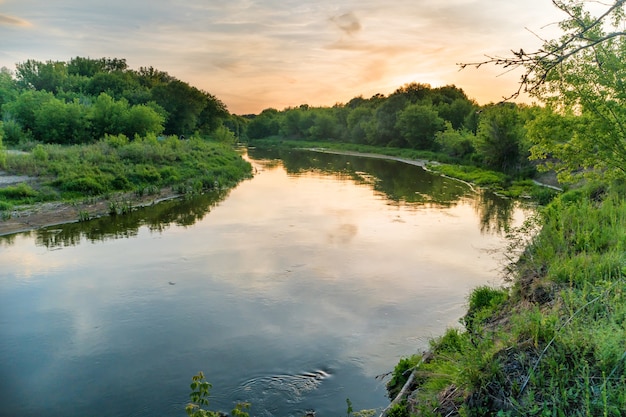 Sonnenuntergang über dem Fluss. Landschaft mit grünen Bäumen, dramatischem Himmel, Wolken und Sonne