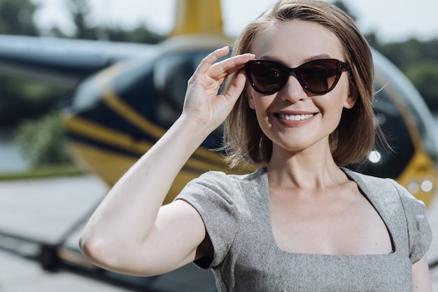 Sonniger Tag. Die Nahaufnahme einer hübschen jungen Geschäftsfrau, die ihre Sonnenbrille anpasst, während sie an einem Hubschrauberlandeplatz posiert