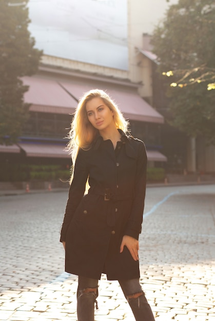 Sonniges Herbstporträt eines fantastischen blonden Models, das schwarzen Mantel trägt und auf der Straße posiert