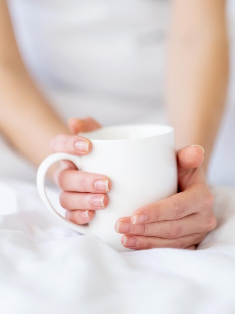 Sonntagskaffee Guten Morgen Entspannung zu Hause Heißes Getränk Nahaufnahme eleganter Frauenhände, die eine Tasse Latte-Getränk auf einer weichen, sauberen weißen Bettlaken-Decke im gemütlichen Bett halten