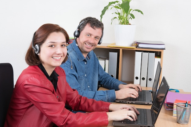 Sonriendo agente de servicio al cliente femenino y masculino usando auriculares con colegas que trabajan en segundo plano en la oficina