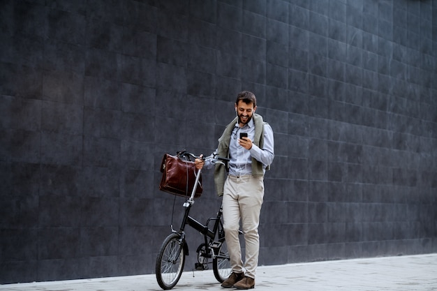 Sonriendo alegre empresario de moda caucásico con teléfono inteligente y empujando su bicicleta. En el fondo es la pared gris.