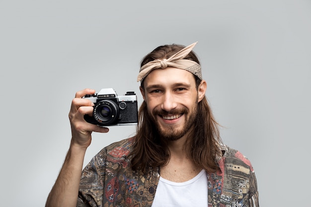 Sonriendo alegre feliz fotógrafo inconformista con una cámara en sus manos fotografías en una cámara de cine, elegantemente vestida con un estilo hippie, persona creativa sobre un fondo blanco de estudio