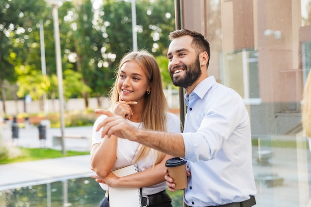 Sonriendo alegre jóvenes colegas de la pareja de negocios con una taza de café y un portátil al aire libre cerca del centro de negocios apuntando a un lado.