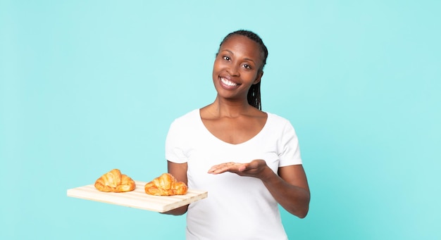 Sonriendo alegremente, sintiéndose feliz y mostrando un concepto y sosteniendo una bandeja de croissant