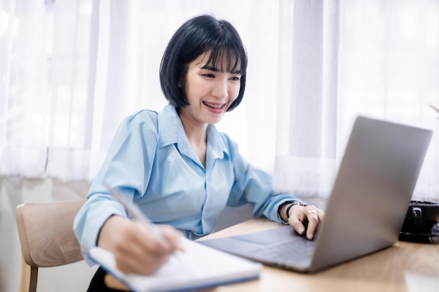 Sonriendo con aparatos dentales de mujeres de negocios asiáticos autónomos Notas escritas en el cuaderno que trabaja con una computadora portátil con una taza de café y un teléfono inteligente en la cafetería