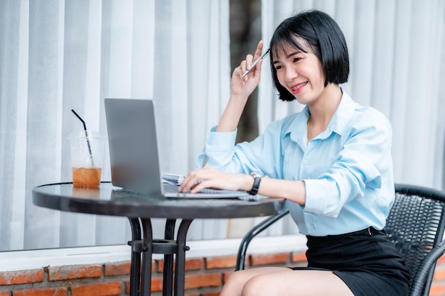 Sonriendo con aparatos dentales de personas asiáticas independientes, mujeres de negocios, toman notas escritas en el teléfono inteligente para trabajar con una computadora portátil con una taza de café y un teléfono inteligente en una cafetería