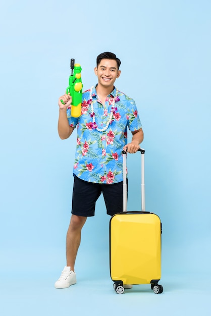 Sonriendo apuesto hombre turista asiático que viaja con pistola de agua y equipaje durante el festival Songkran