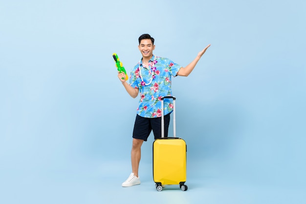 Sonriendo apuesto hombre turista asiático que viaja con pistola de agua y equipaje durante Songkran