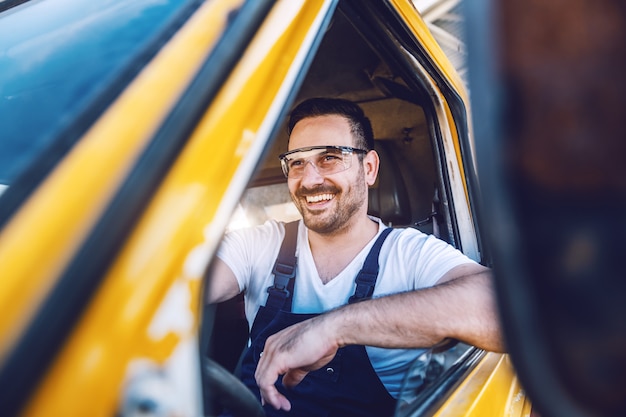 Sonriendo apuesto trabajador sin afeitar conducir vehículo en sitio de construcción.