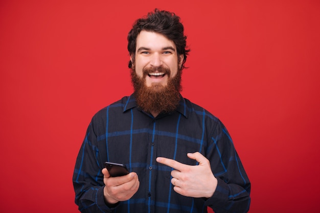 Sonriendo barbudo apuntando al móvil en la mano, mientras que sobre la pared roja