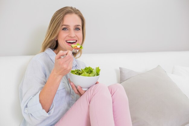 Sonriendo casual rubia sentada en el sofá comiendo ensalada